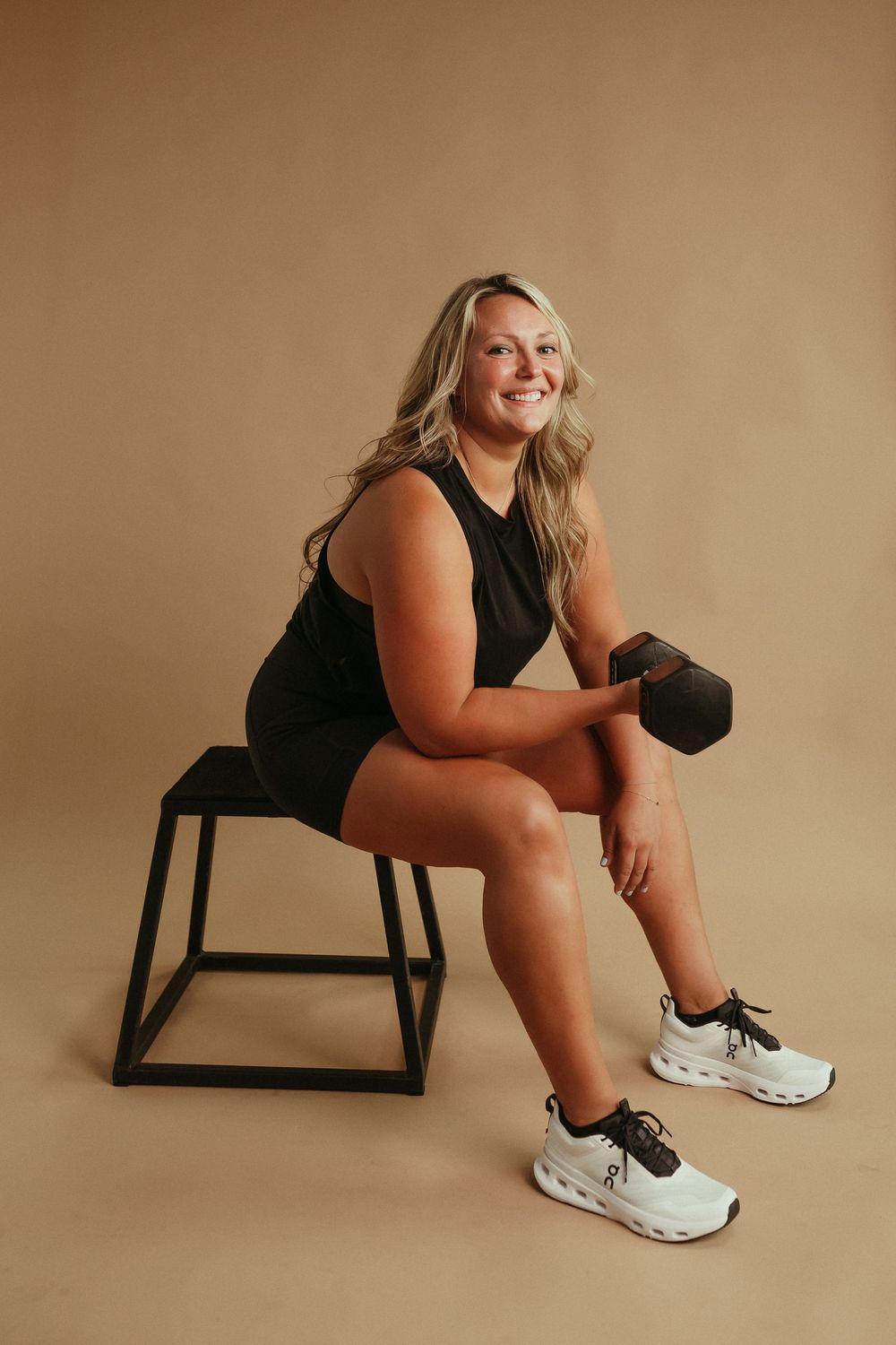 Woman in black athletic wear sits on bench holding dumbbell during fitness photoshoot with beige background.
