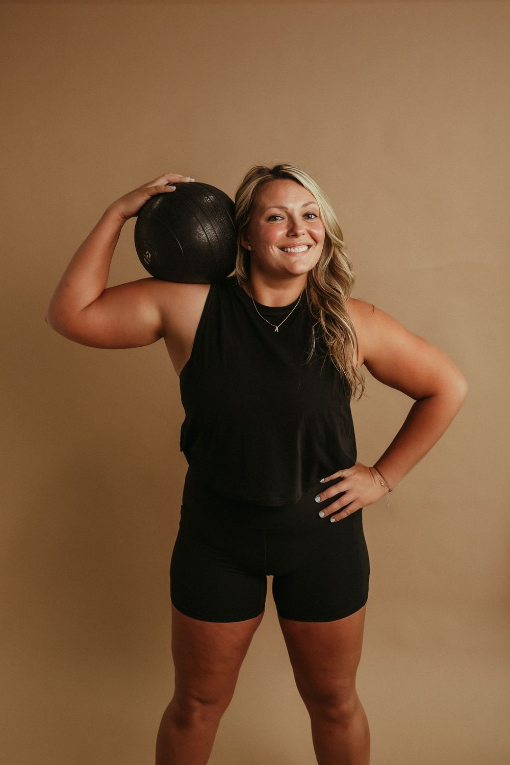 Athletic model in black workout attire holds medicine ball while posing confidently against brown backdrop.