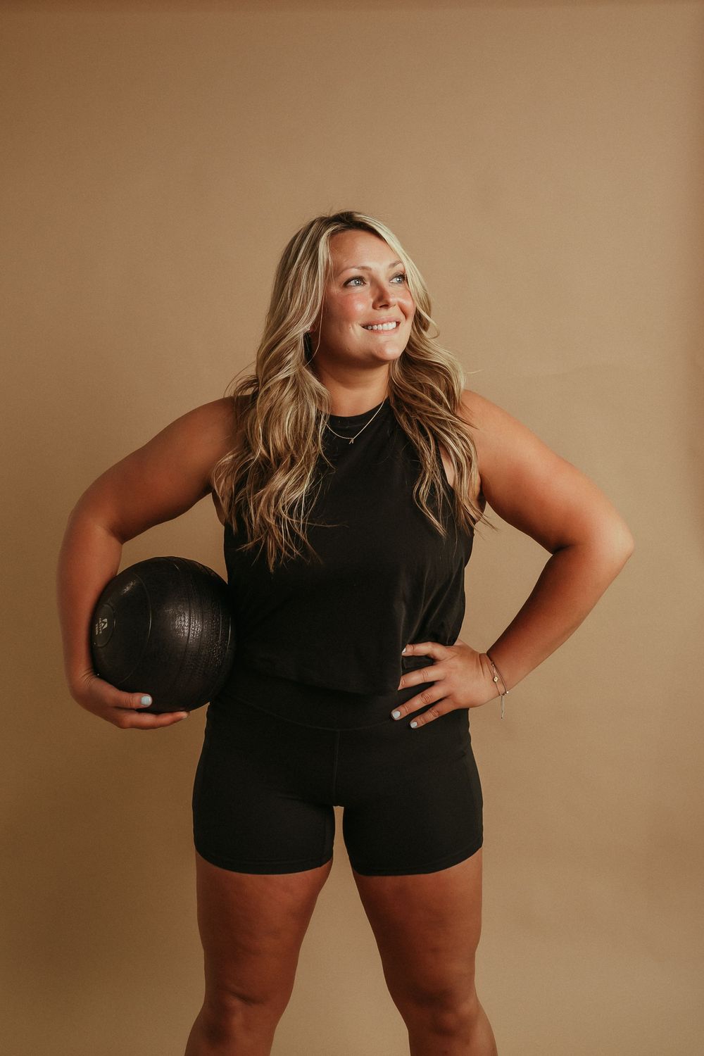Confident fitness model in black athletic wear holds medicine ball against neutral studio background.