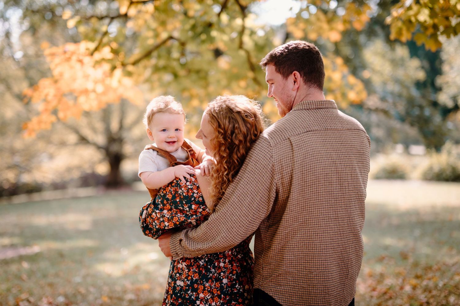 A couple embraces outdoors in a park during autumn with golden leaves visible in the background.