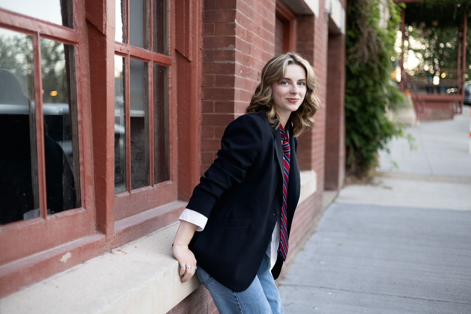 A stylish figure in jeans and a dark jacket poses along an urban brick building sidewalk.