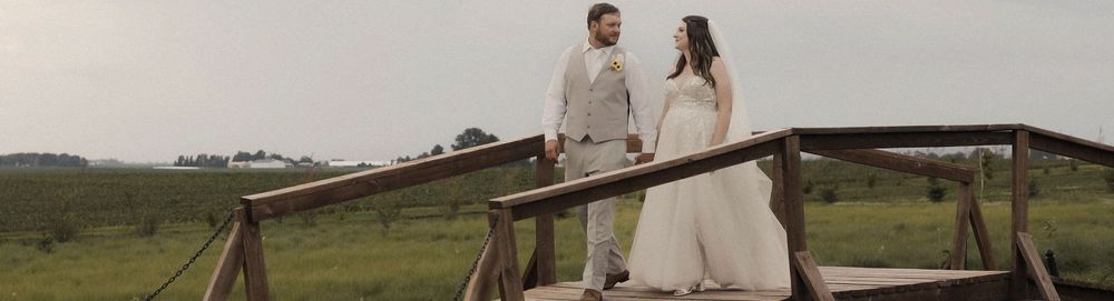 A couple stands on a wooden bridge in a scenic rural field with stormy skies in the background.