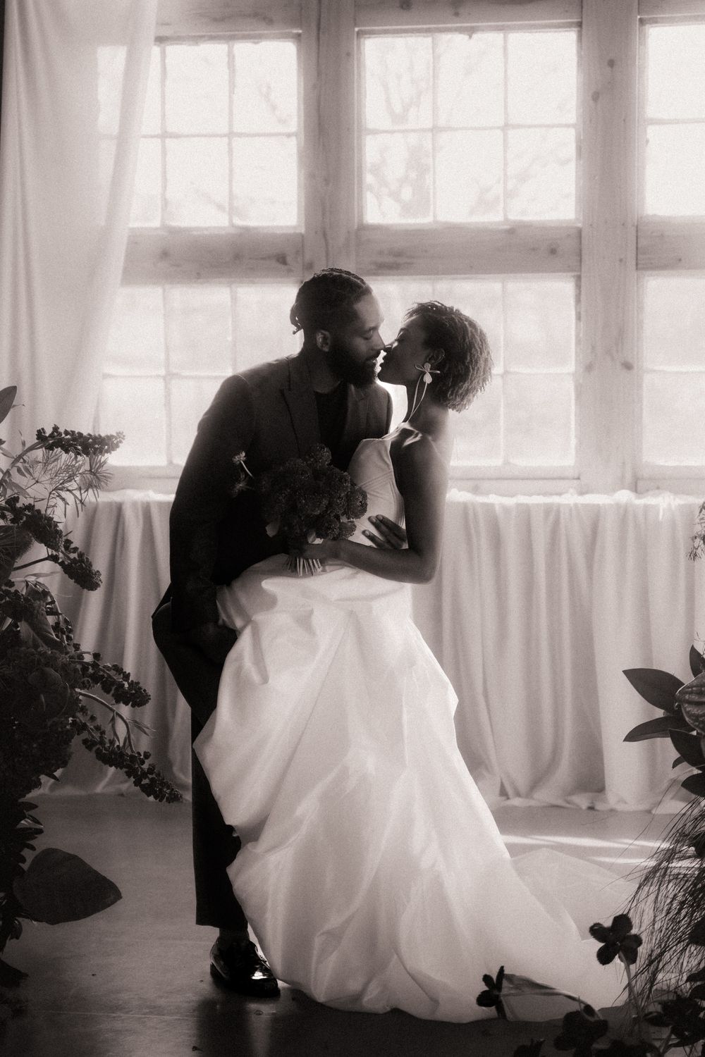 A romantic wedding moment silhouetted against bright windows, featuring a bride in white gown and groom sharing an intimate moment.