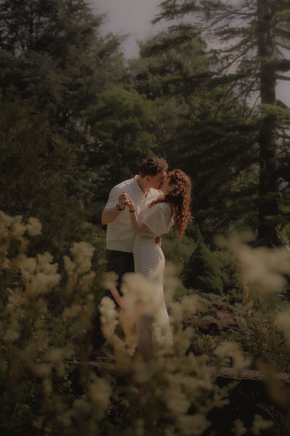 Romantic couple embraces in moody forest setting with soft lighting and wildflowers in foreground.