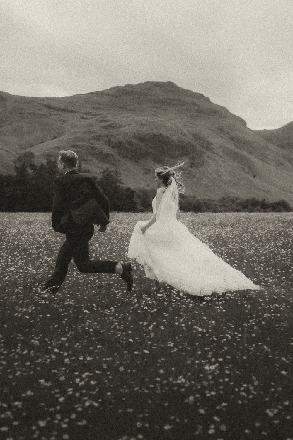 A couple in wedding attire runs through a picturesque meadow filled with wildflowers against a backdrop of rolling hills.