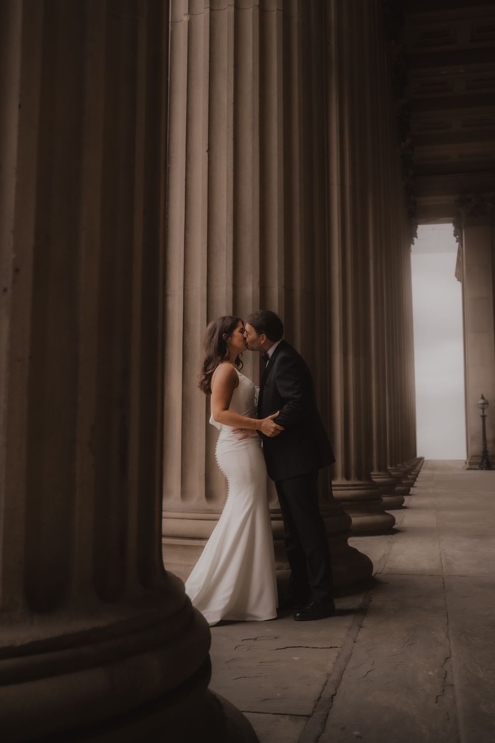 Romantic wedding couple embraces between dramatic stone columns in a moody architectural setting at dusk.