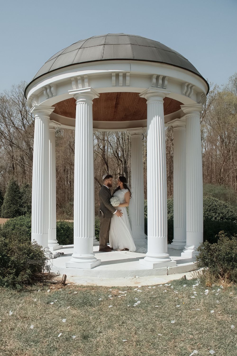 A couple embraces in an elegant white gazebo with Corinthian columns surrounded by trees and greenery.