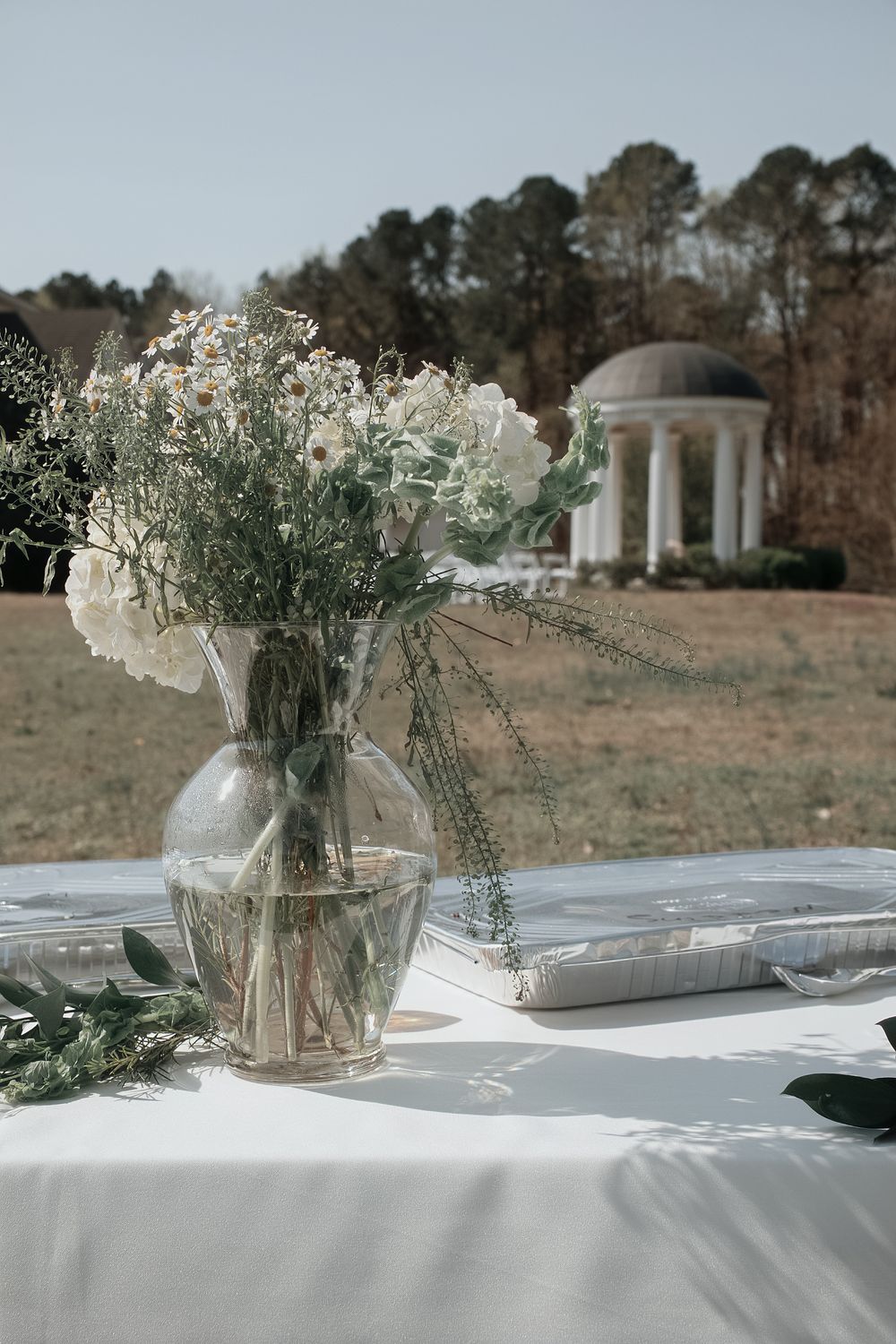 White floral centerpiece in glass vase on table with gazebo in background at outdoor garden venue.