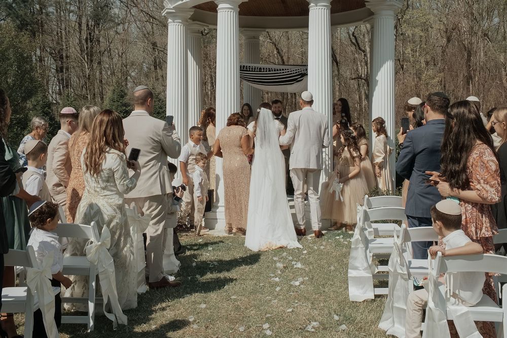 Outdoor wedding ceremony under a white gazebo with guests seated on white chairs surrounded by early spring trees.