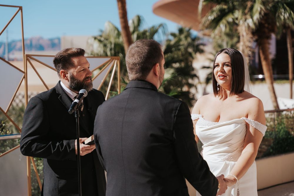 A wedding ceremony takes place outdoors with palm trees in the background on a sunny day.