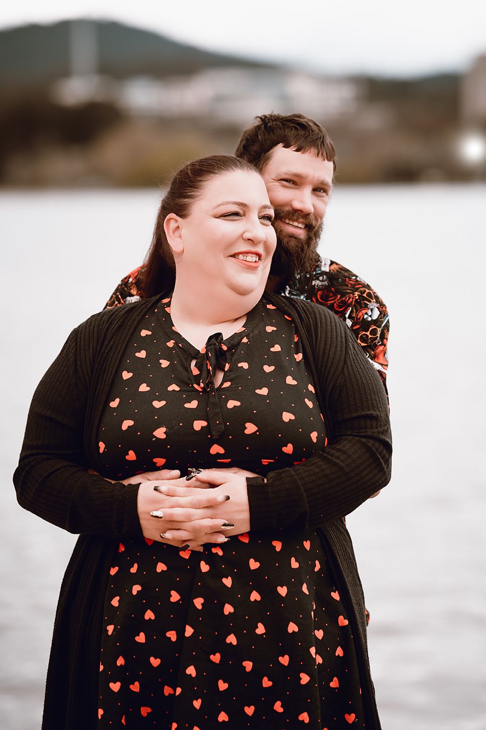Couple embraces outdoors by water, wearing matching dark clothing with heart print dress against blurred nature background.