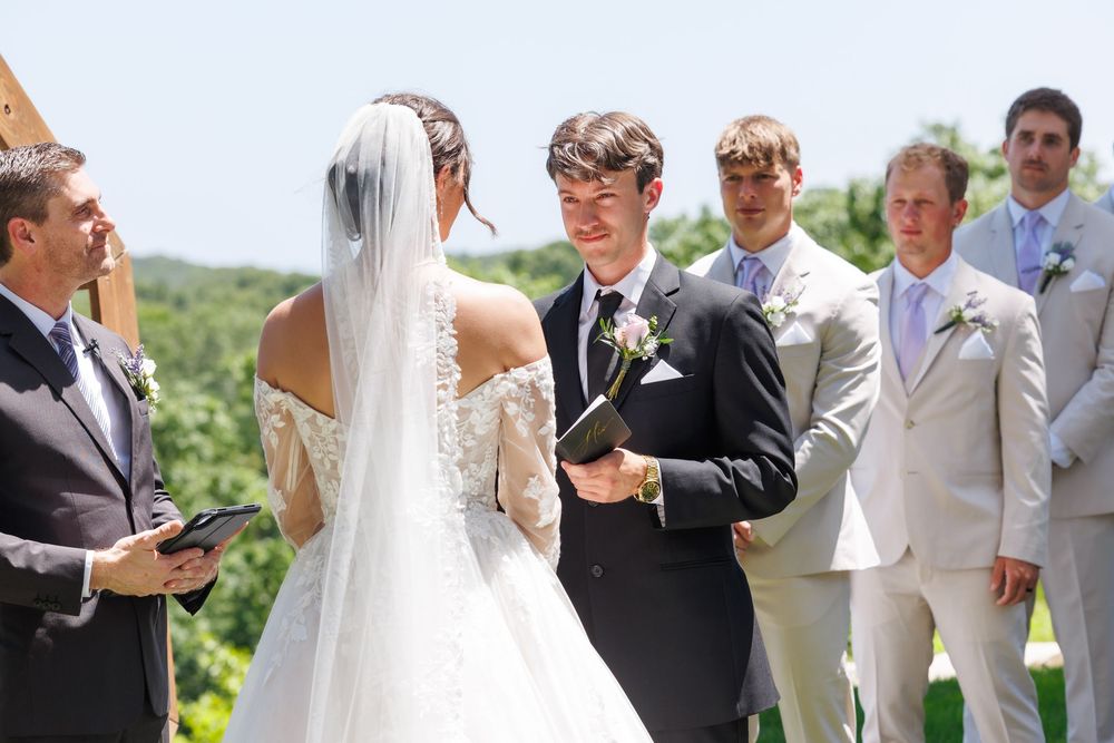 Outdoor wedding ceremony with bride in white dress and veil exchanging vows with groom surrounded by wedding party.