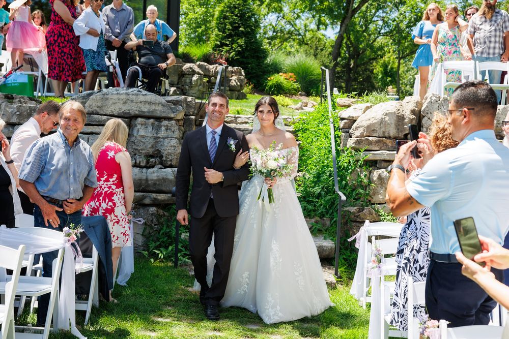 A garden wedding ceremony takes place outdoors with guests seated on white chairs amidst lush greenery.