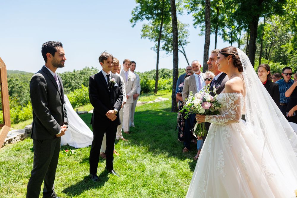Wedding ceremony taking place outdoors on a sunny day with green trees and grass in the background.