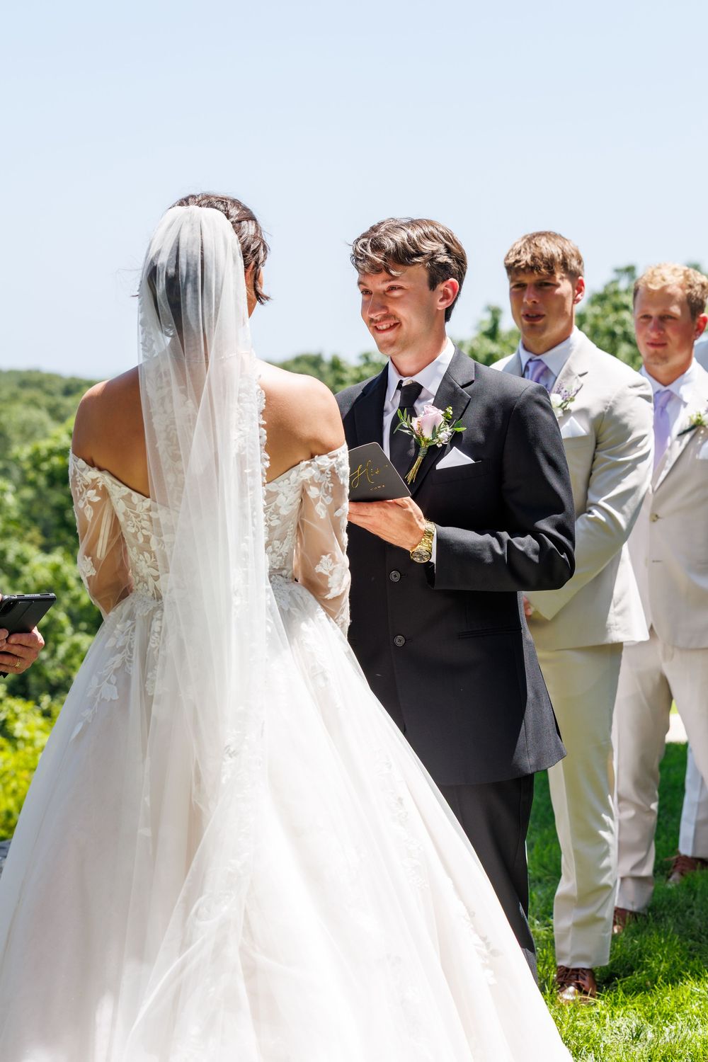 A wedding ceremony takes place outdoors on a sunny day with the bride in a flowing white dress and guests gathered.