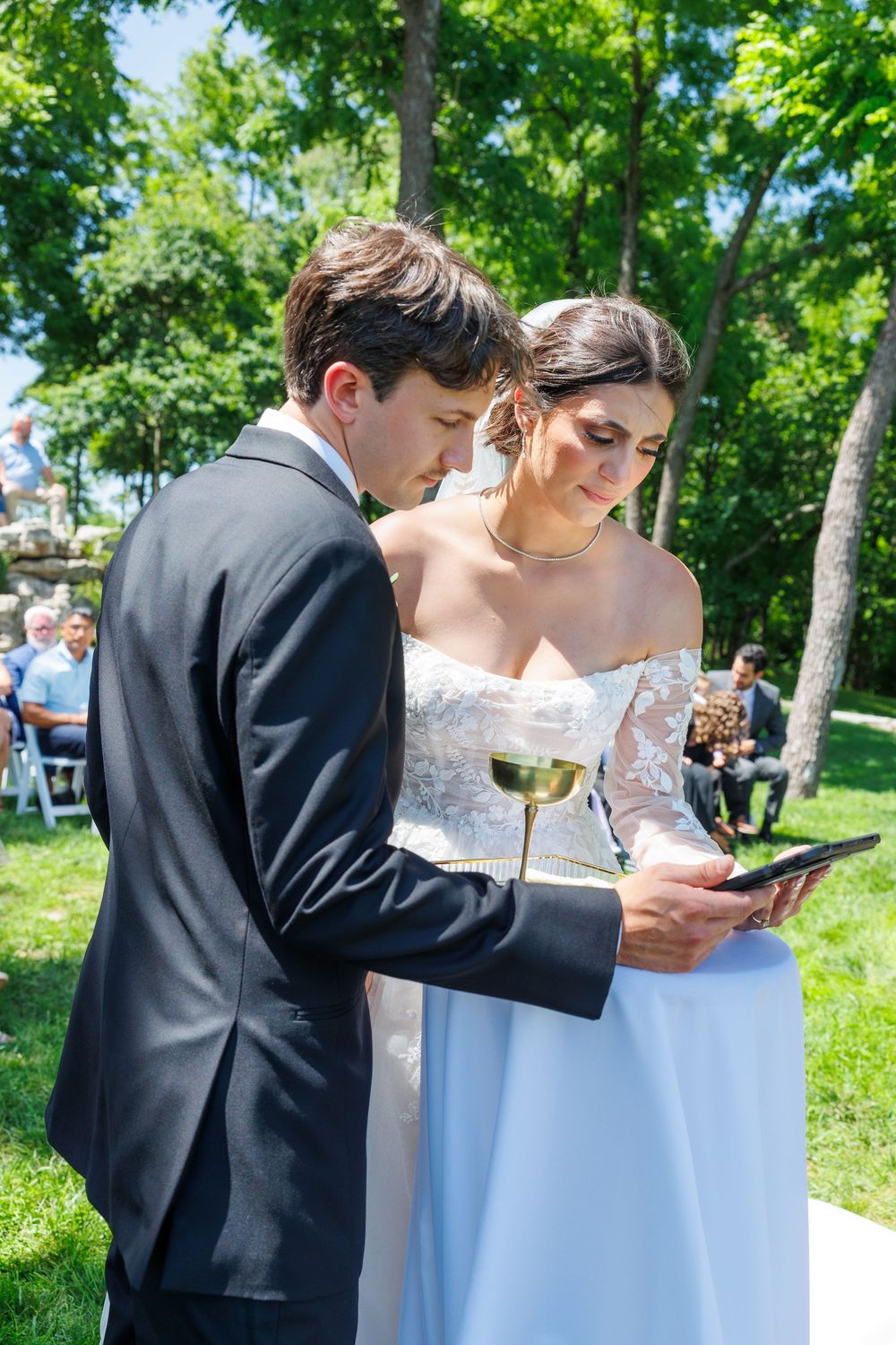 Couple signing documents outdoors at summer garden wedding ceremony with guests in background.