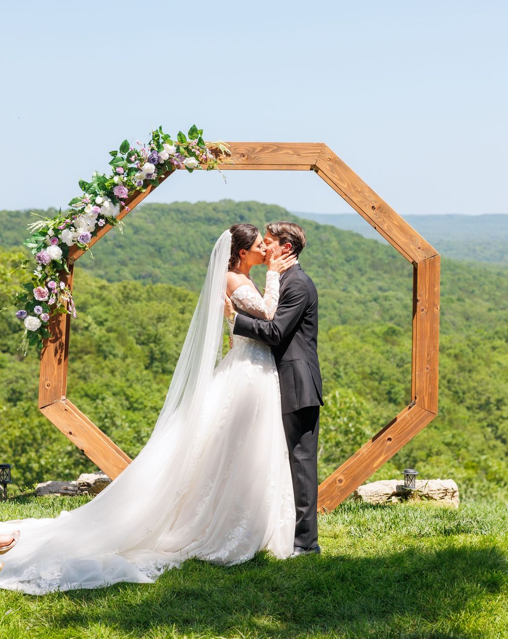 A couple shares a romantic moment under a wooden octagonal arch decorated with flowers against a scenic mountain backdrop.