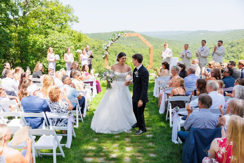 Outdoor wedding ceremony with guests seated in white chairs on a hilltop overlooking a scenic valley landscape.