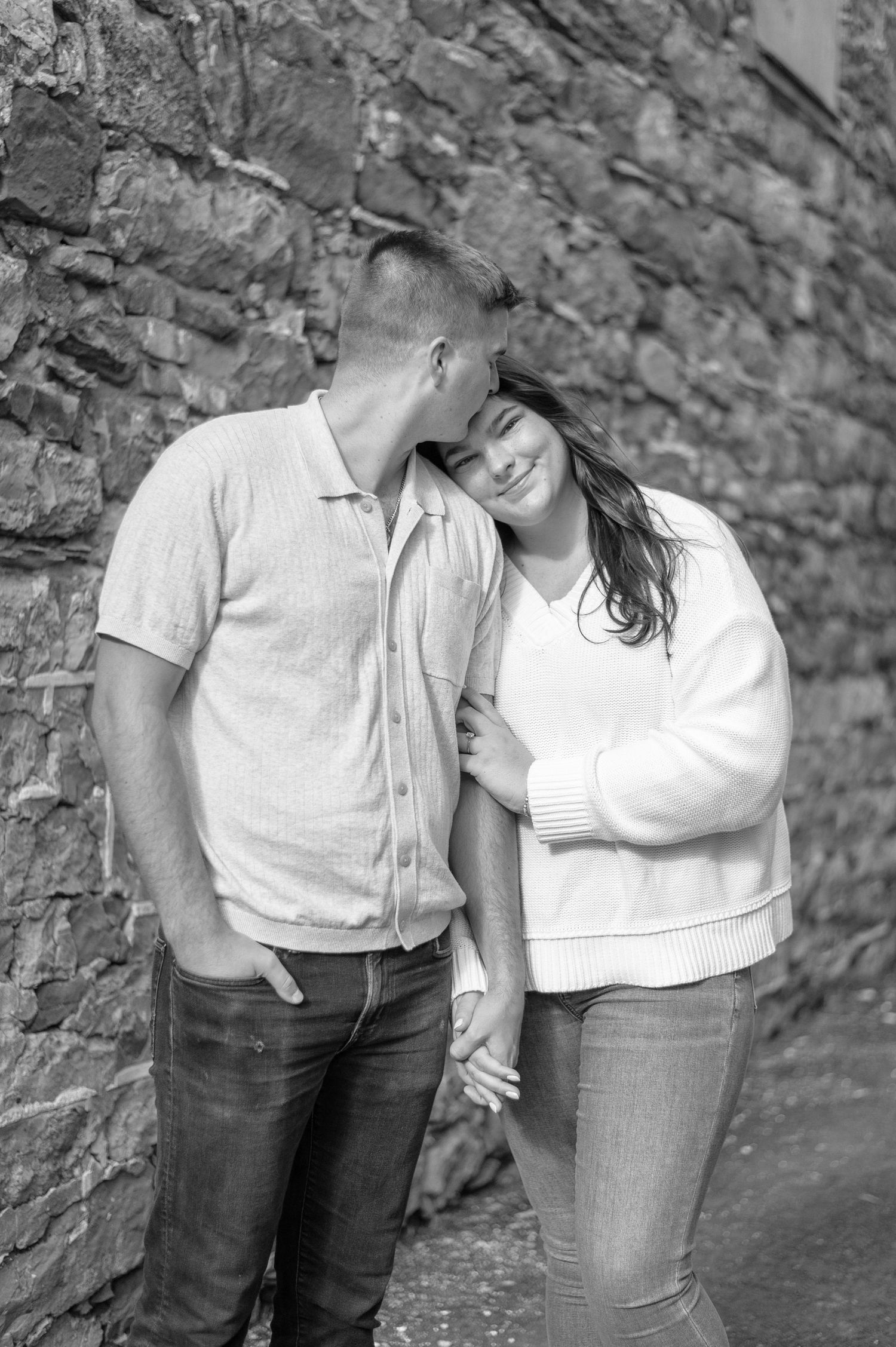 A romantic black and white photo of a couple leaning against a stone wall.