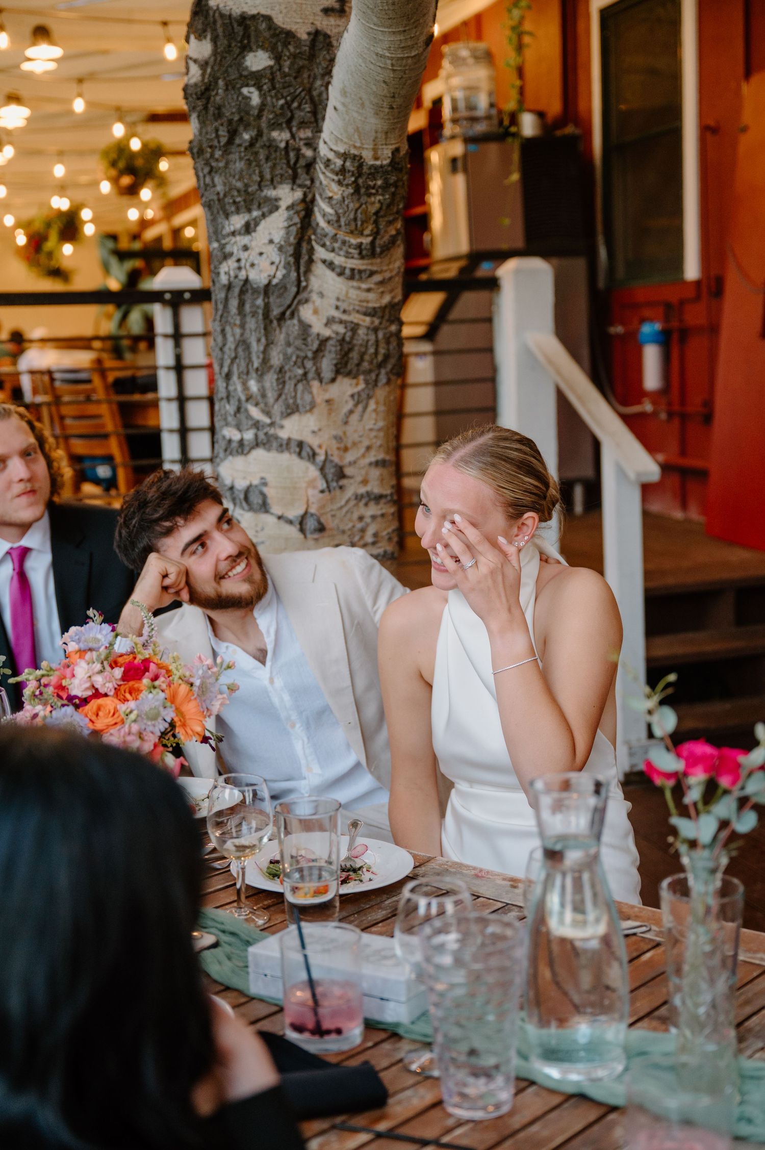 A couple shares a candid moment at an outdoor restaurant table decorated with flowers and string lights.