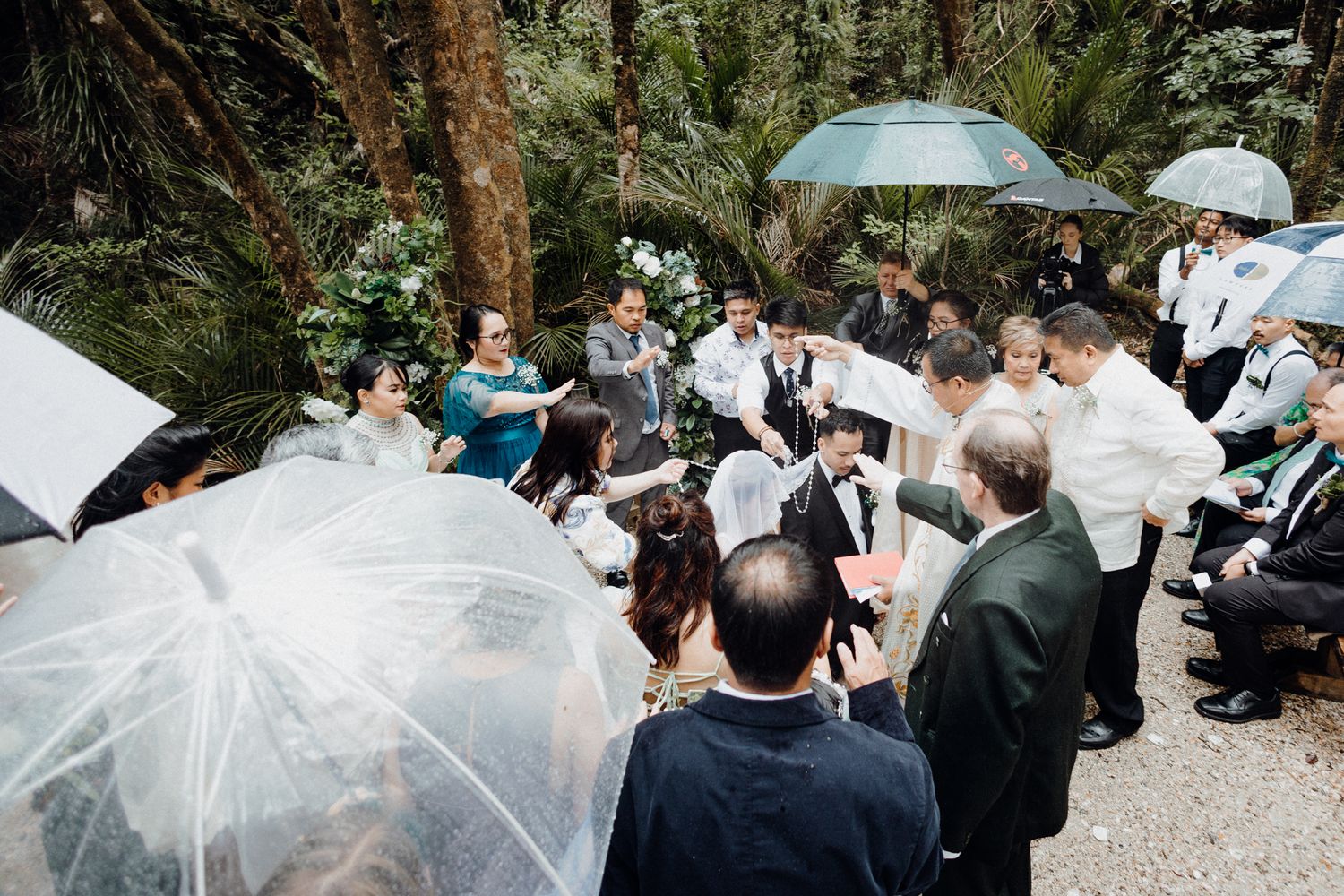 A crowd gathers with umbrellas for an outdoor
wedding ceremony captured by Apertured Media Auckland Wedding Photographer.