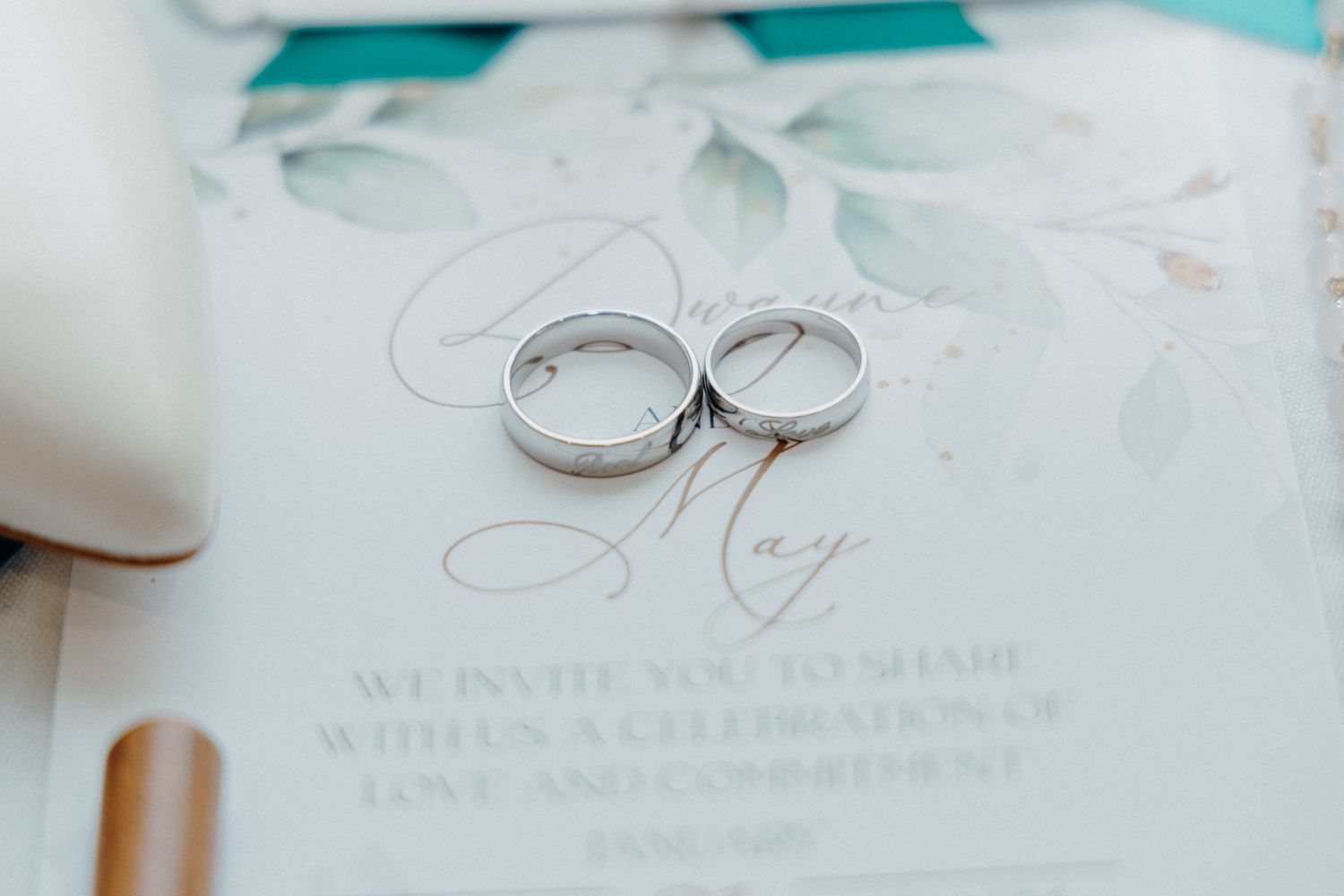 Two silver wedding rings rest on a wedding invitation with elegant cursive captured by Apertured Media Auckland Wedding Photographer.