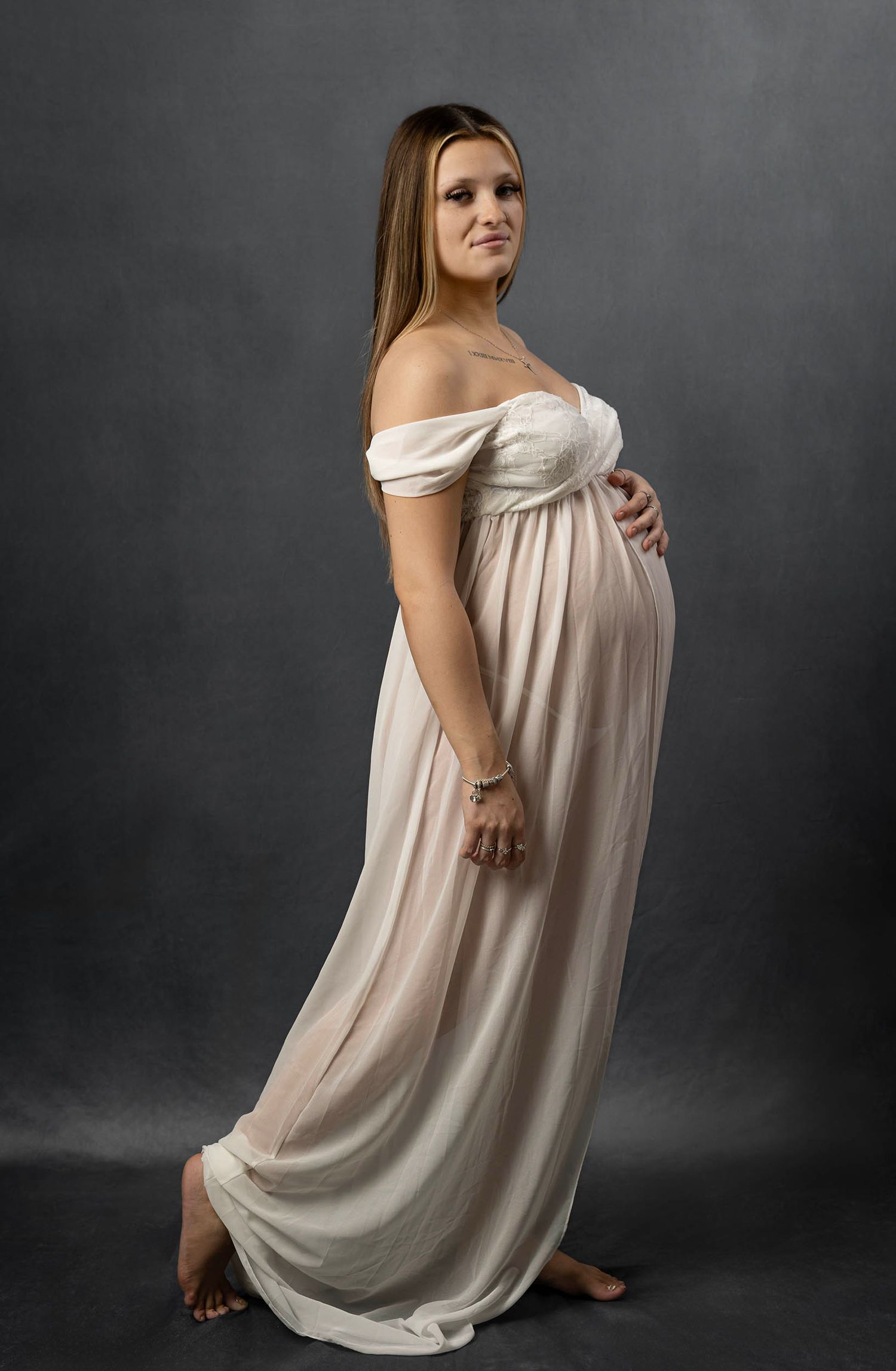 Elegant maternity portrait in cream gown against a dark gray background. Maternity photo shoot, pregnancy photography Hampshire studio.