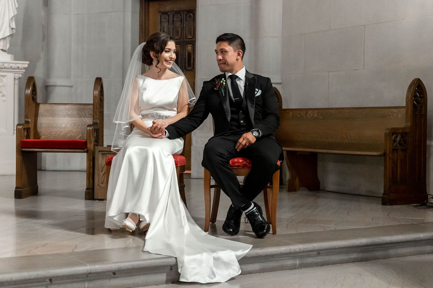 Wedding couple shares a candid moment while seated on wooden chairs in an elegant church sanctuary.