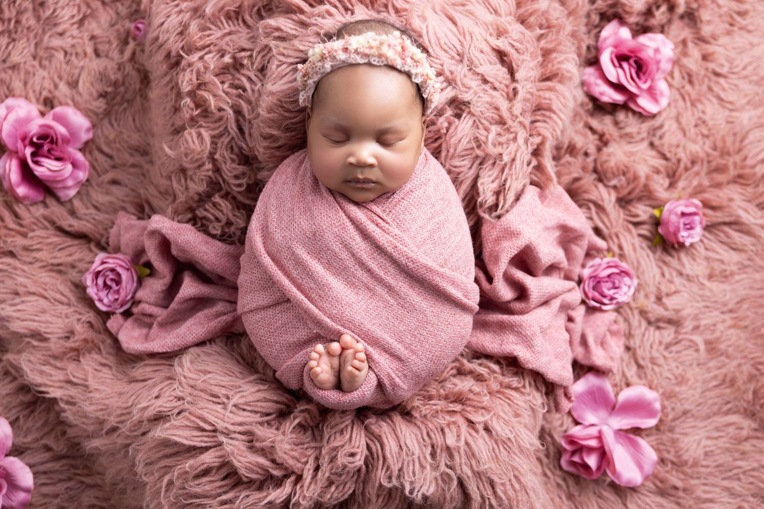 A sleeping newborn swaddled in pink fabric lies on fluffy pink fur surrounded by matching pink roses.