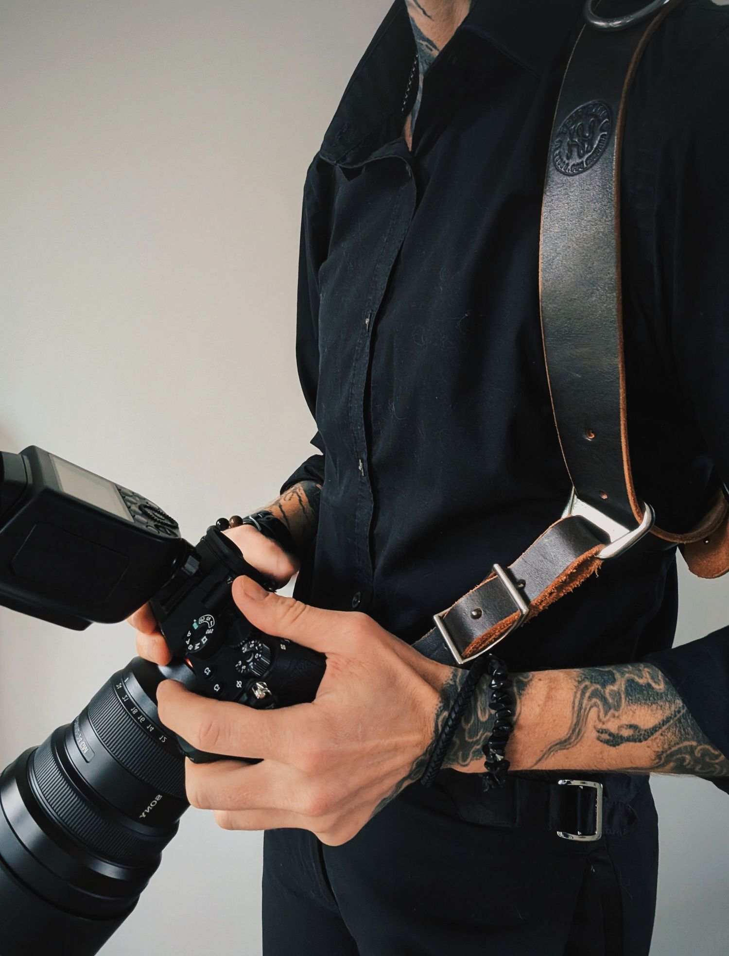 Close-up of hands holding professional camera equipment with visible tattoos and leather straps.