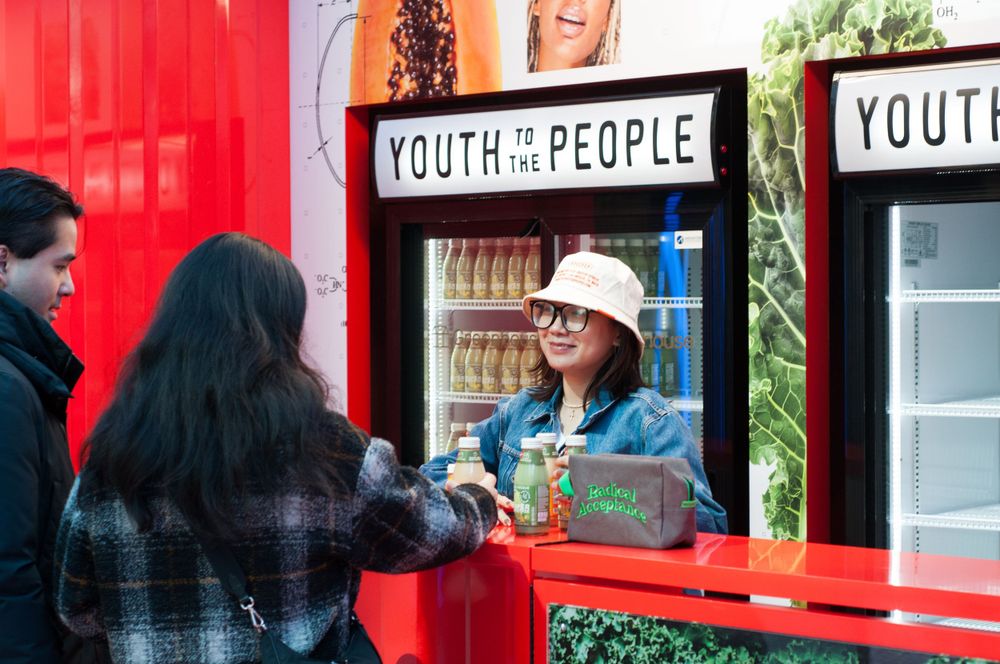 A retail worker helps customers at a bright red Youth to the People skincare kiosk or storefront counter.