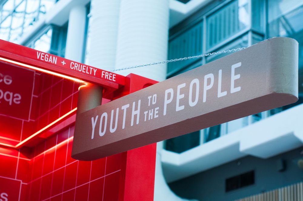 A modern street sign reading 'Youth to the People' glows with red light against an aqua blue building facade.