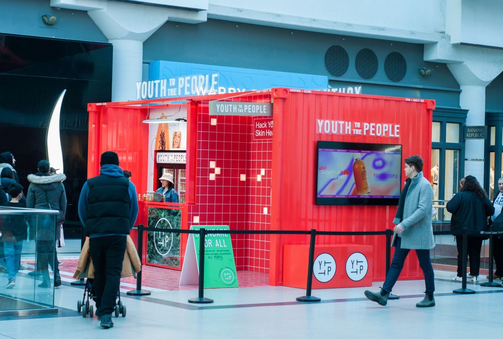 A bright red retail kiosk with digital displays and mosaic tiles stands in front of a blue building while people walk past.