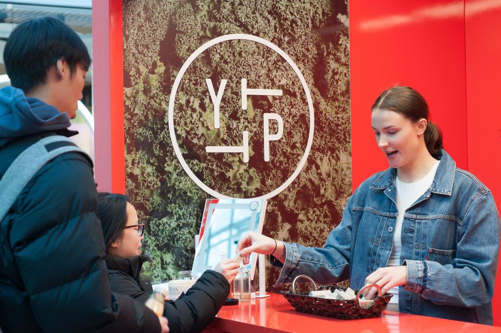 People interact at a coffee shop counter with a YP logo visible in the background on a red wall.