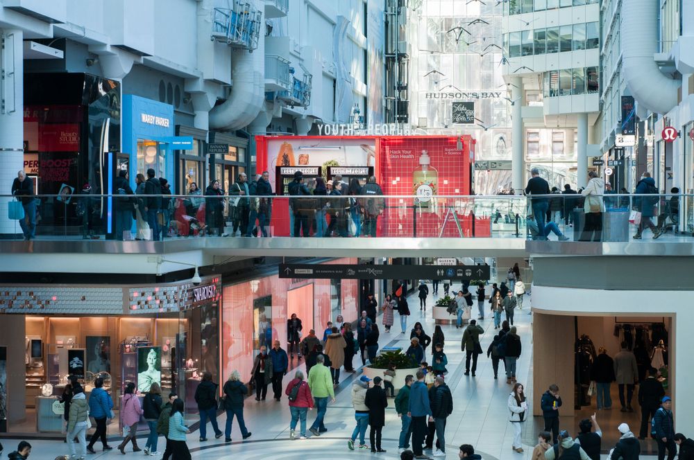Busy shopping mall interior with multiple levels showing shoppers walking between retail stores with bright signage.
