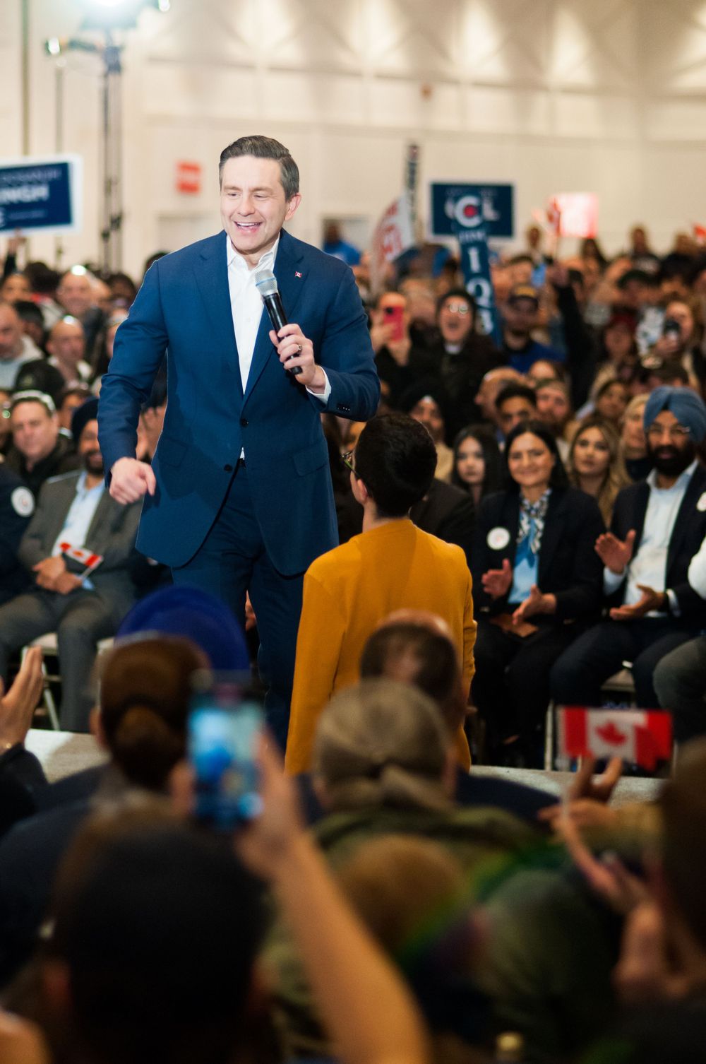 A speaker in a navy suit addresses a large crowd at a campaign rally as attendees hold phones and signs in the audience.