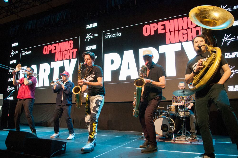 Musicians performing energetically on stage with brass instruments and a large illuminated PARTY sign display in the background.