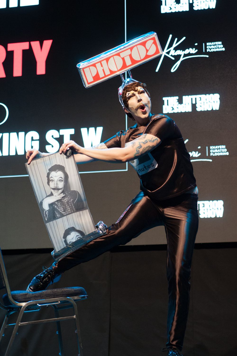 Performer in black outfit strikes dramatic pose on stage while holding a framed photograph during show presentation.