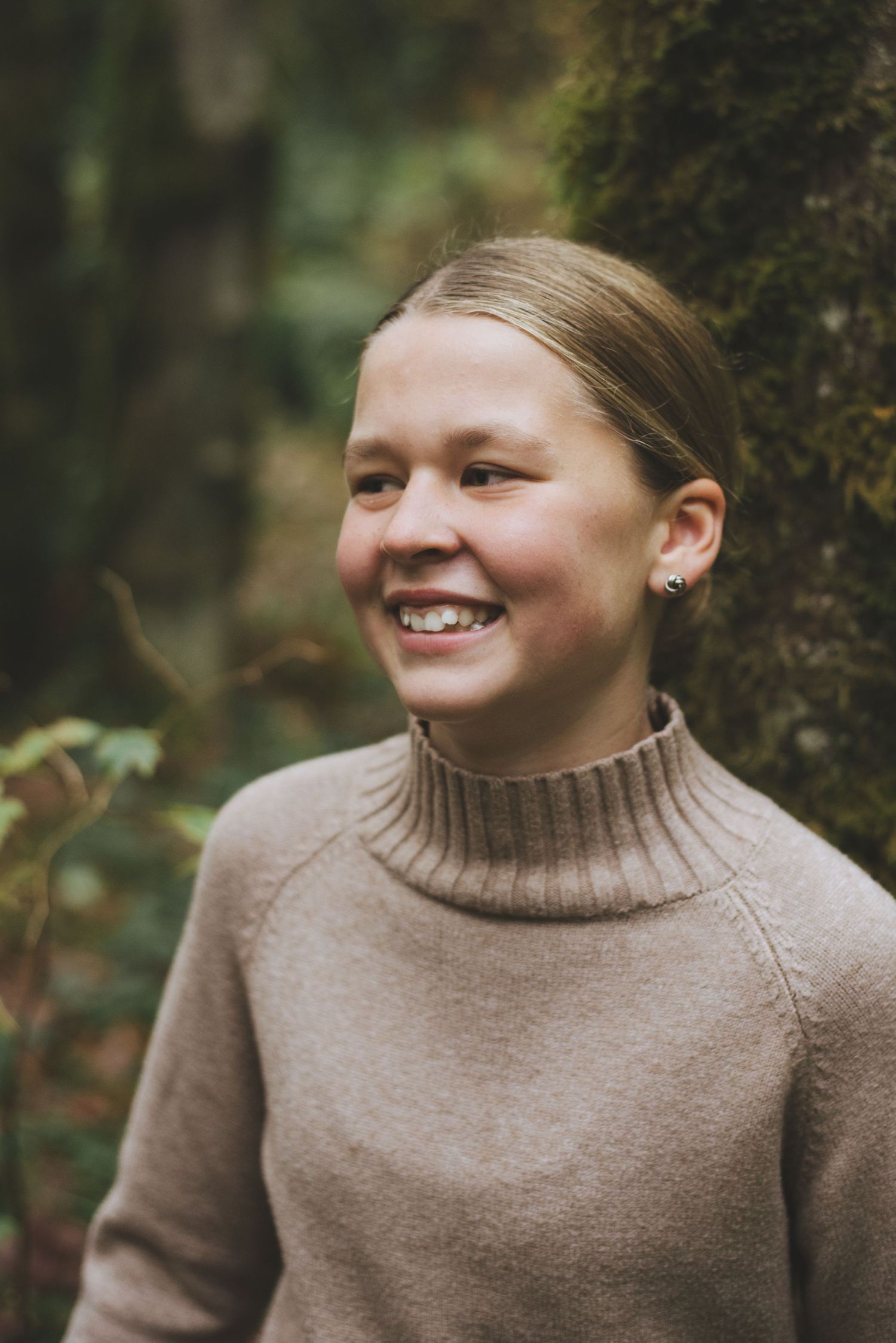 Teenaged girl in beige sweater smiles while posing for photos in the woods at goldstream provincial park.