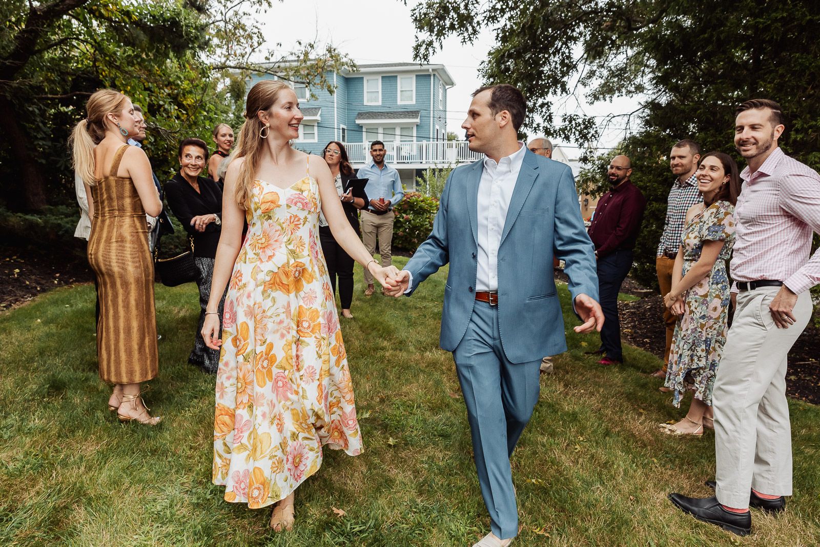 A couple walks together at an outdoor gathering, with the woman wearing a floral dress and man in a blue suit.