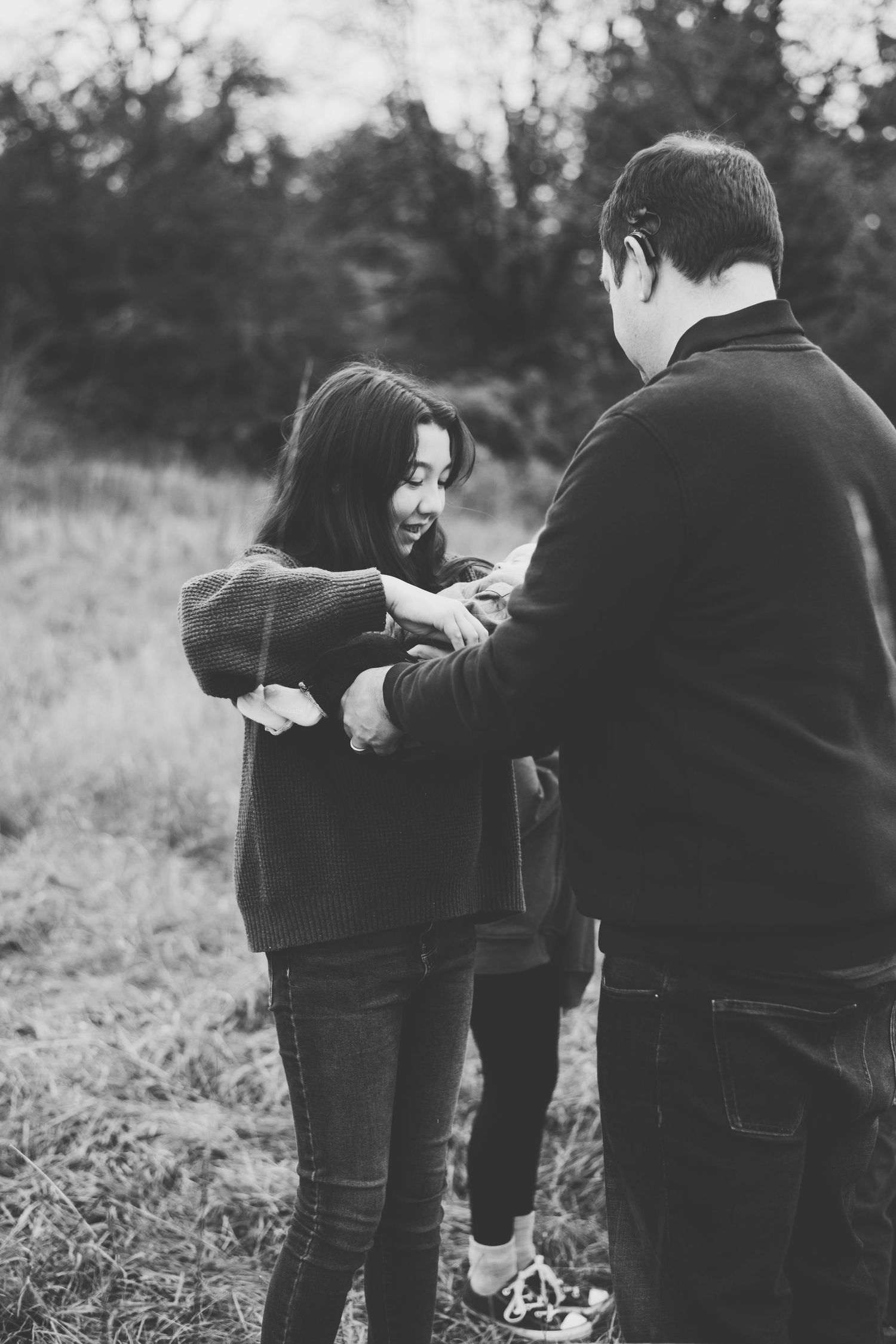 A father hands a baby to his teenaged daughter outdoors in a grassy field.