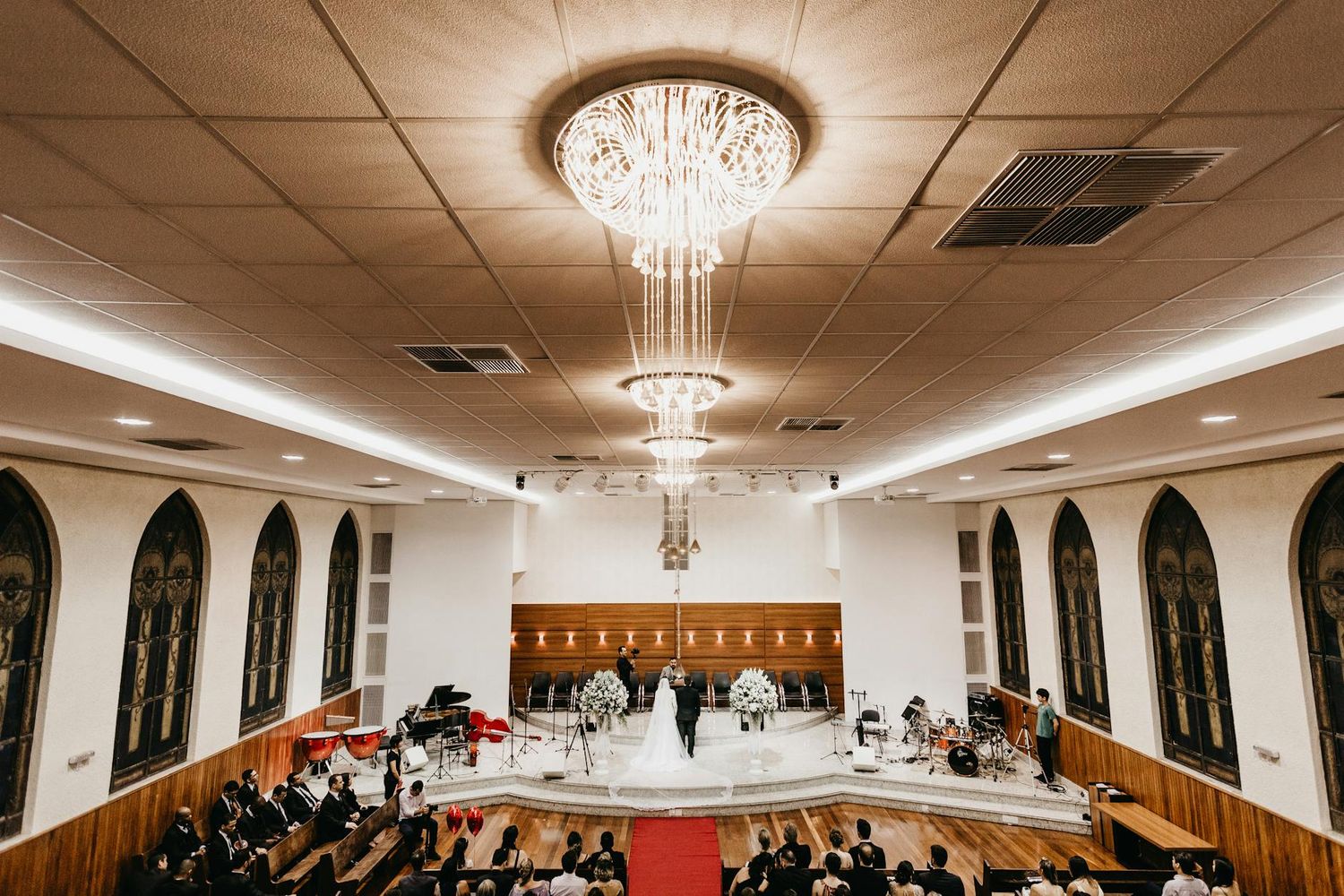 Interior view of an elegant wedding ceremony venue with arched windows, crystal chandeliers, and guests seated in rows.