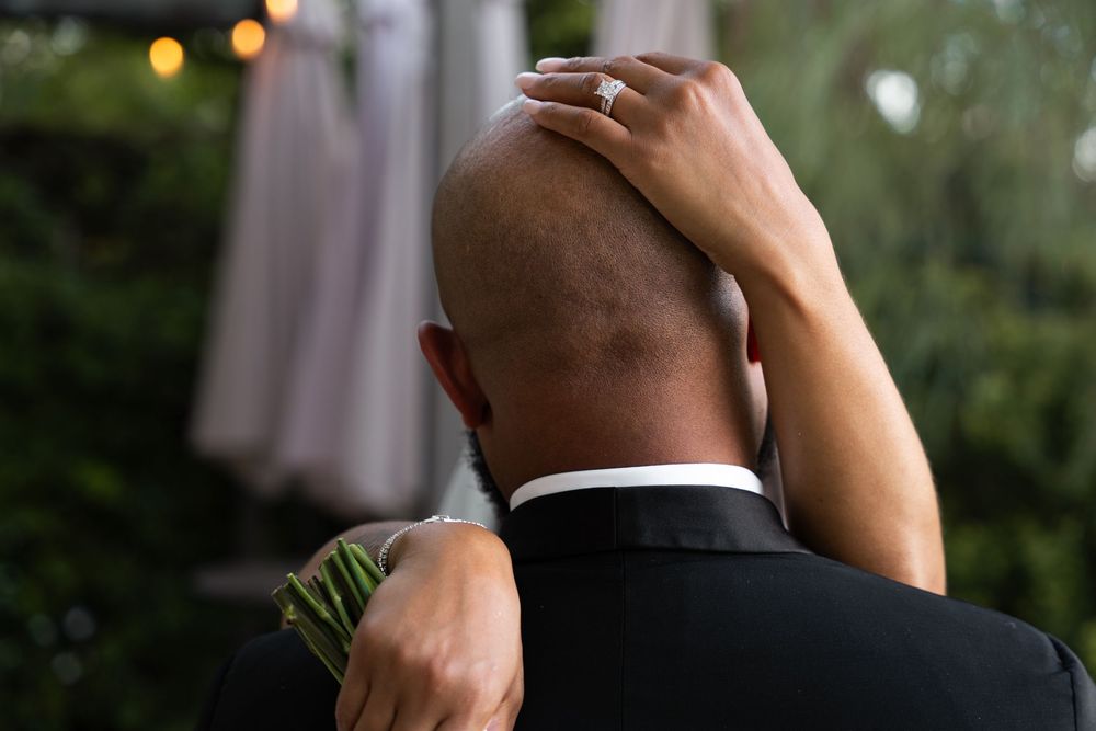 A person in a black suit holds their head from behind while standing outdoors at dusk with bokeh lights in the background.