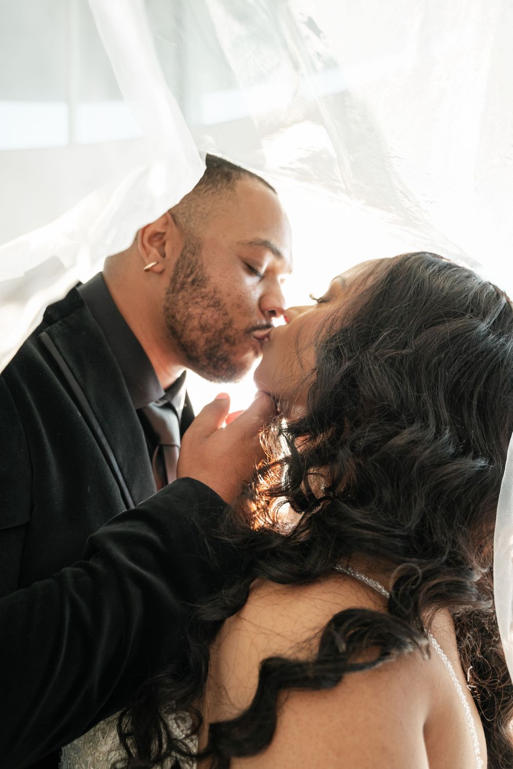 An intimate romantic moment between a couple sharing a tender kiss amidst sheer white curtains in soft light.