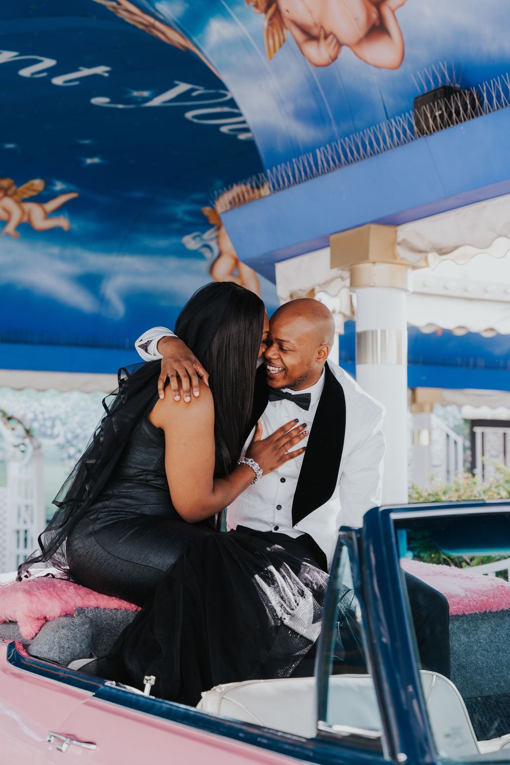 A romantic moment captured of a couple in formal attire sitting in a classic convertible car beneath a blue sky.