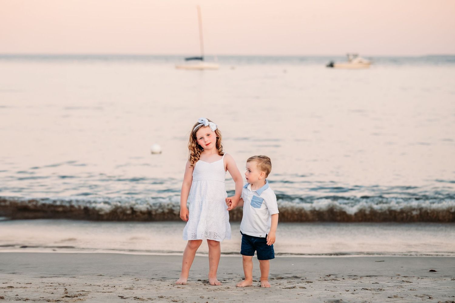 Two young children in light-colored outfits stand together at the beach during sunset with waves rolling in behind them.