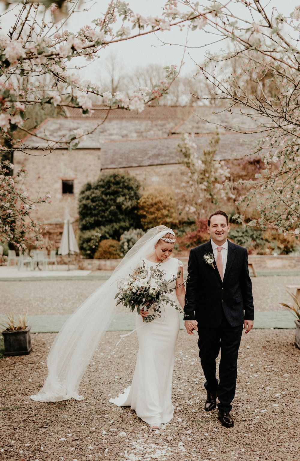 A couple on their wedding day walk together in a garden with warm autumn colors and flowering trees in the background.