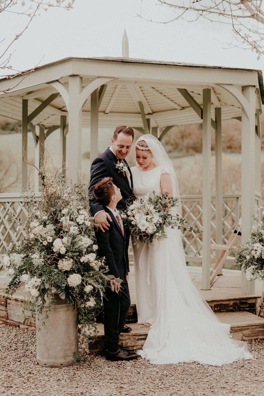 A romantic wedding ceremony takes place under a white wooden gazebo decorated with elegant floral arrangements.
