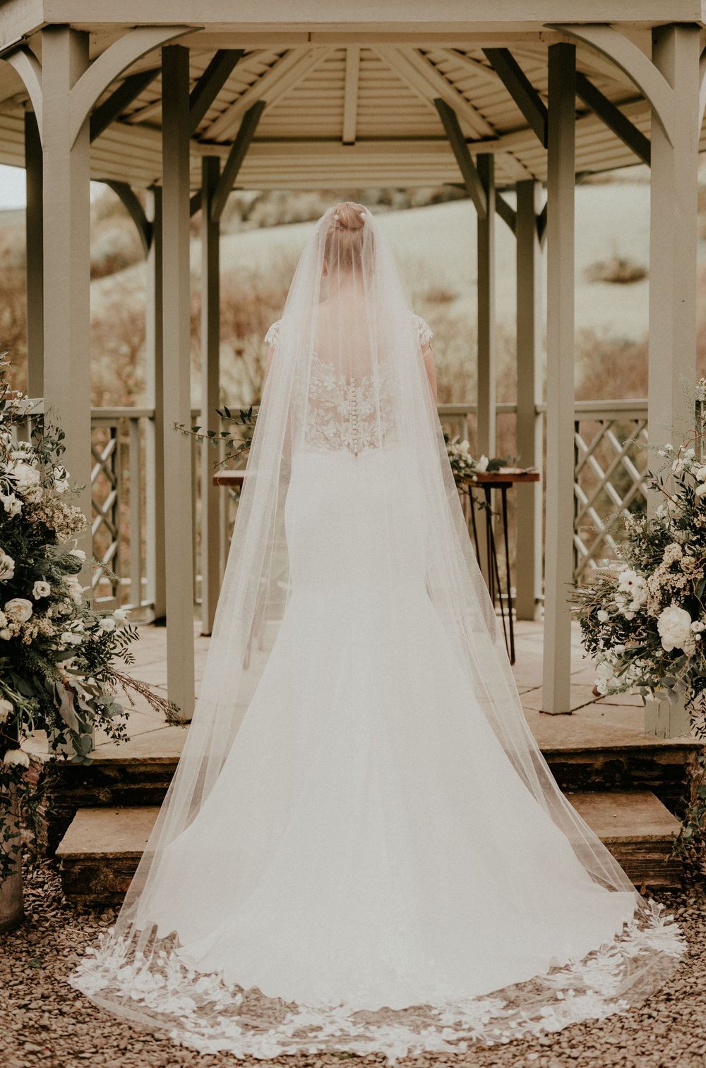 A long white wedding dress and veil photographed from behind in a wooden gazebo decorated with floral arrangements.