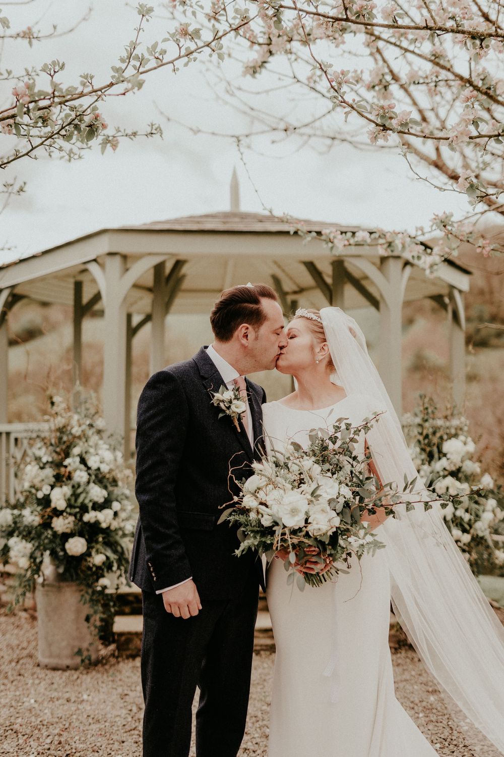 A romantic wedding kiss underneath a white gazebo with blooming spring trees and floral arrangements in the background.